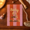 1367pkbabyitscold Avayves Feine Papeterie Und Geschenke Für Babys Und Kids Greeting card on a striped pink/orange background that says 'Baby it’s cold outside' on a wooden table.