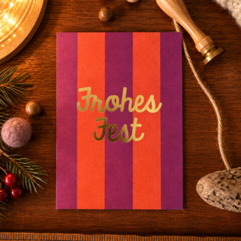Card with orange and purple vertical stripes and gold 'Frohes Fest' text on a wooden table with holiday decor