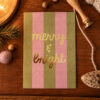 Holiday card with pink and olive green vertical stripes and gold foil text reading 'merry & bright' on a wooden desk surrounded by seasonal props.