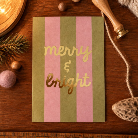 Holiday card with pink and olive green vertical stripes and gold foil text reading 'merry & bright' on a wooden desk surrounded by seasonal props.