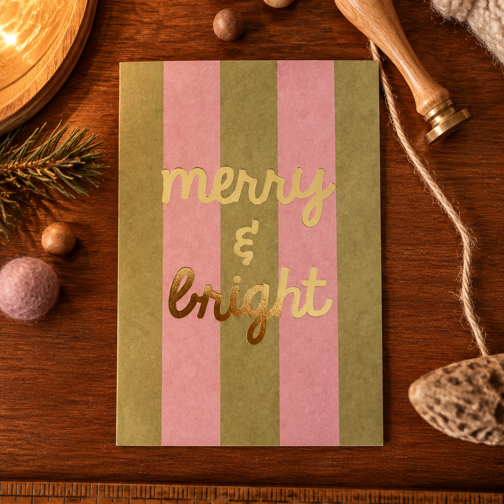 Holiday card with pink and olive green vertical stripes and gold foil text reading 'merry & bright' on a wooden desk surrounded by seasonal props.