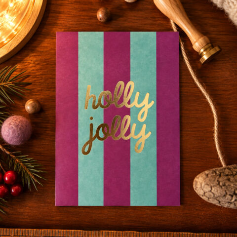 Festive card with purple and teal vertical stripes and gold 'holly jolly' text on a wooden table, surrounded by Christmas decor.