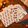 4138adventcalendarhaeuser02 Avayves Feine Papeterie Und Geschenke Für Babys Und Kids Assorted Christmas gift wrap papers in red, blue, and cream patterns spread on a wooden table with fairy lights.