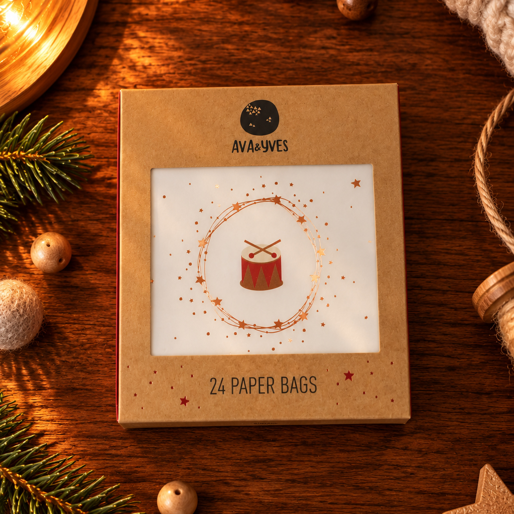 Brown kraft packaging for 'AVA&YVES' 24 paper bags, with a circular star motif and red drum logo shown through a clear window, set on a wooden table with holiday decorations around it.