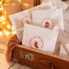 Vintage brown suitcase filled with white holiday cards featuring red stocking and house stamps, glowing fairy lights in the background.