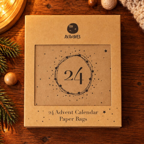 Brown cardboard box labeled '24 Advent Calendar Paper Bags' with a circular logo, on a wooden table with pine branches and warm string lights.