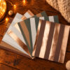 Color swatch cards with copper metallic stripes fanned on a wooden table with warm fairy lights and a knitted blanket nearby.
