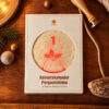 White Advent calendar box on a wooden table; round window shows the number 1 with a red holly design around it. Elements around include pine, string, and a ruler edge in the frame.
