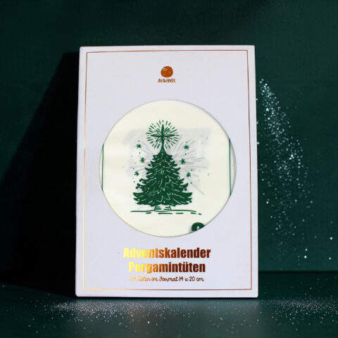 White Advent calendar box with a round window showing a green Christmas tree design and gold 'Adventskalender' text below, festive packaging edge visible.
