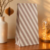 Tall diagonal-striped gift bag on a wooden table, with a ceramic vase on the left and a glass cloche with warm fairy lights on the right nearby.