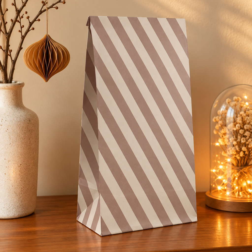 Tall diagonal-striped gift bag on a wooden table, with a ceramic vase on the left and a glass cloche with warm fairy lights on the right nearby.