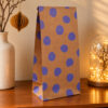 5537papierbeuteldots Avayves Feine Papeterie Und Geschenke Für Babys Und Kids Purple-dotted brown paper gift bag on a wooden table with warm lighting and decorative vase nearby.