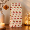 5539papierbeutelhaeuser Avayves Feine Papeterie Und Geschenke Für Babys Und Kids Tall gift bag with red houses pattern on beige background sits on a wooden table next to a lit candle and a decorative glass dome with lights.