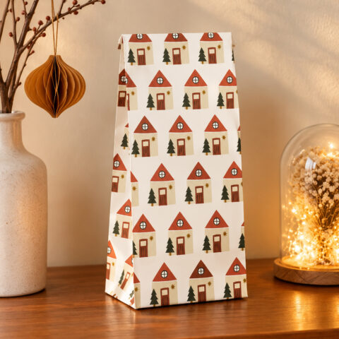 Gift bag with a repeating red-roofed houses pattern on a wooden table, beside a ceramic vase and a glass dome with warm fairy lights.