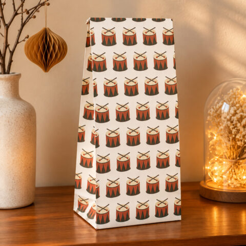 Tall white gift bag with a repeating red drum motif on a wooden table, flanked by a speckled vase on the left and a glass cloche with dried flowers on the right.