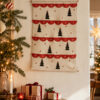 Advent calendar with red numbered doors and small Christmas trees on a beige wall, beside a wooden sideboard with gifts and a lit candle.