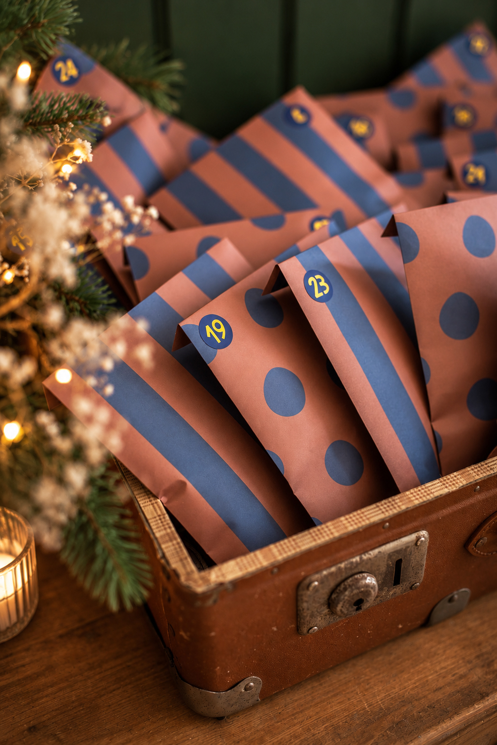 Brown vintage suitcase filled with pink and blue polka-dotted Advent calendar bags labeled with numbers, set under warm Christmas lights.