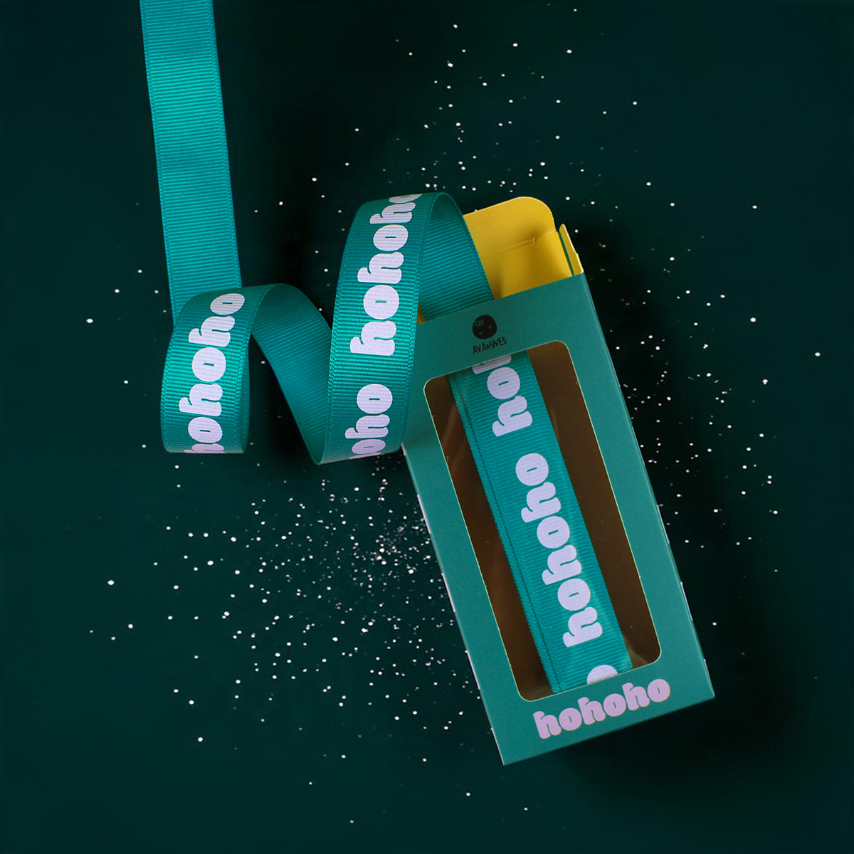 Teal gift box with a teal ribbon reading 'hohoho' looping around a yellow-inside box on a dark green background with white confetti specks.