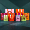 Colorful gift bags of various sizes with ribbon handles arranged on a dark teal surface, festive prints visible.