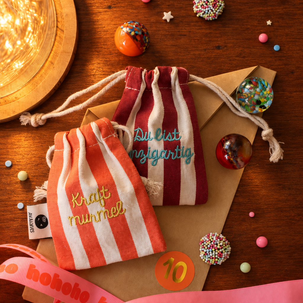Two striped fabric drawstring pouches on a wooden table beside a gift envelope, candy decorations, and a pink ribbon; embroidered text visible on the pouches.|