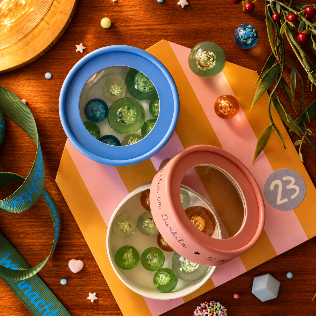 Top-down view of two round party containers with blue and pink rims, filled with colorful glass marbles, on a striped orange‑pink card surrounded by confetti and ribbons.
