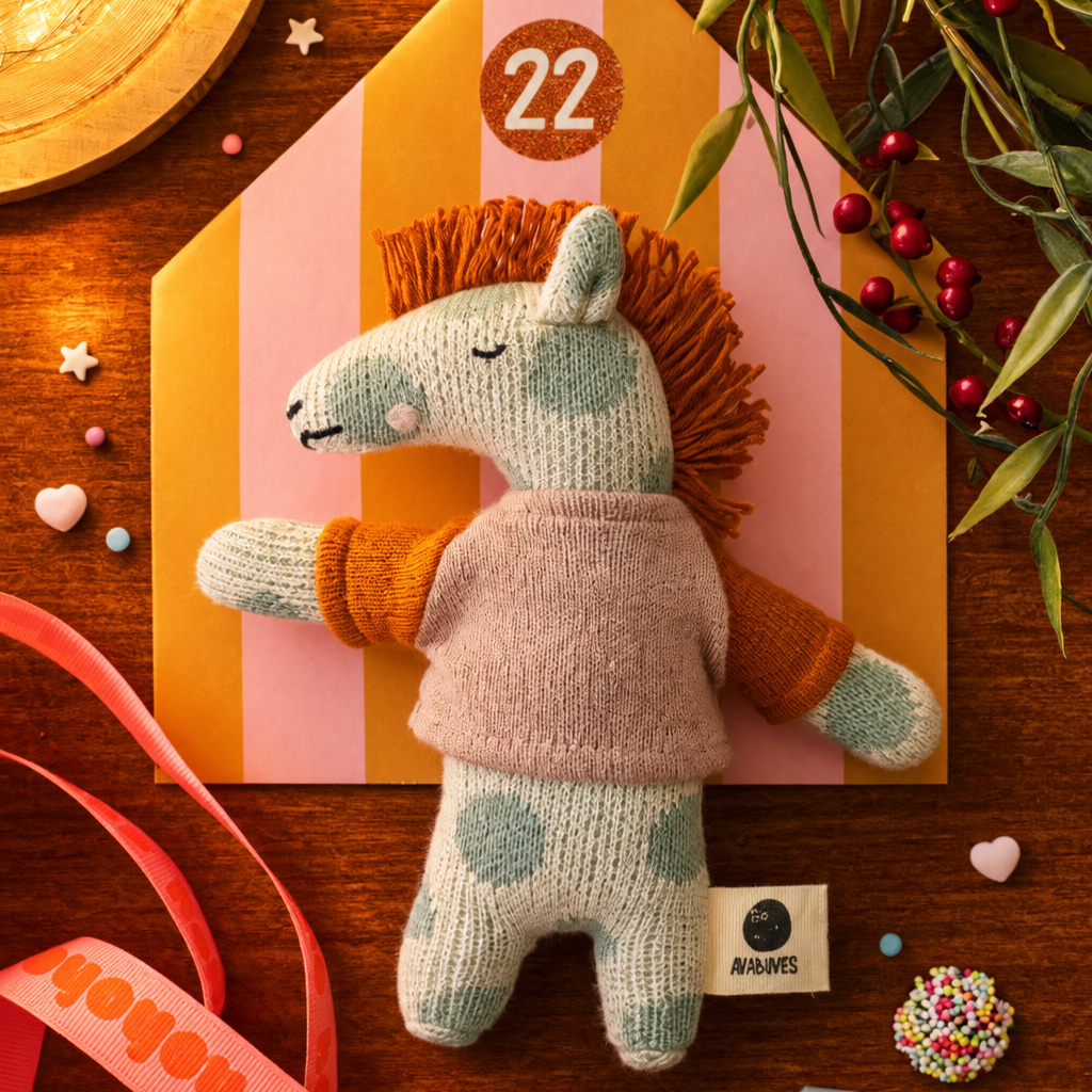 Soft knitted horse plush with orange mane on a pink-and-orange striped advent calendar backdrop marked 22, surrounded by ribbons and festive decorations.