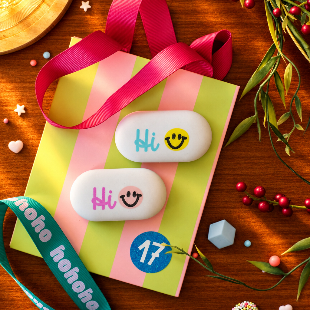 Two white oval soaps with 'Hi' and a smiley face rest on a pastel-striped gift bag with pink ribbon, in a festive flat lay.