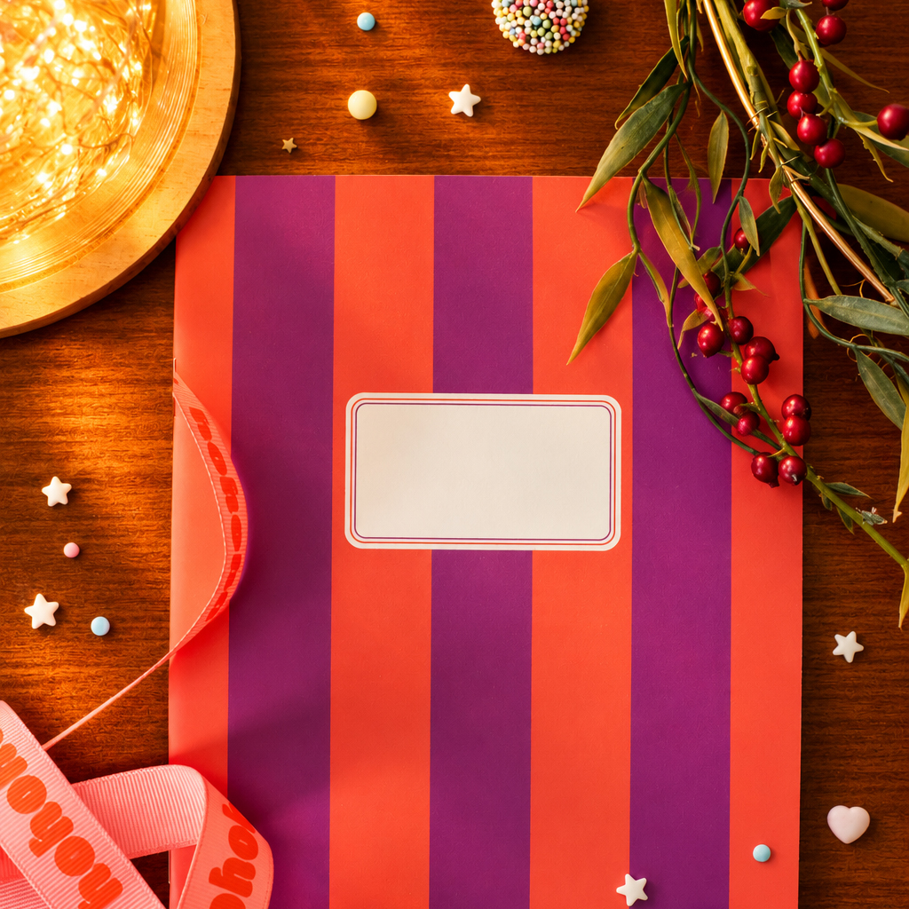 Gift bag with purple and orange stripes on a wooden table, blank label on front, surrounded by festive decorations like ribbon and berries.