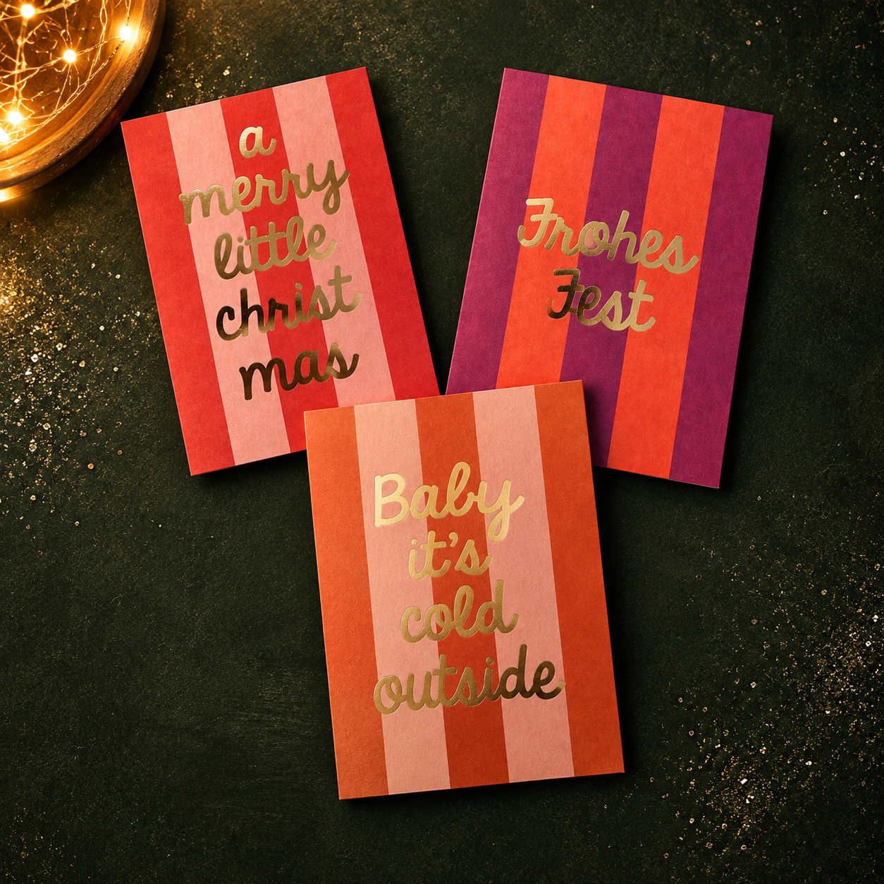 Three striped holiday cards with gold script text: 'a merry little christmas', 'Frohes Fest', and 'Baby it's cold outside' on a dark surface with warm fairy lights.