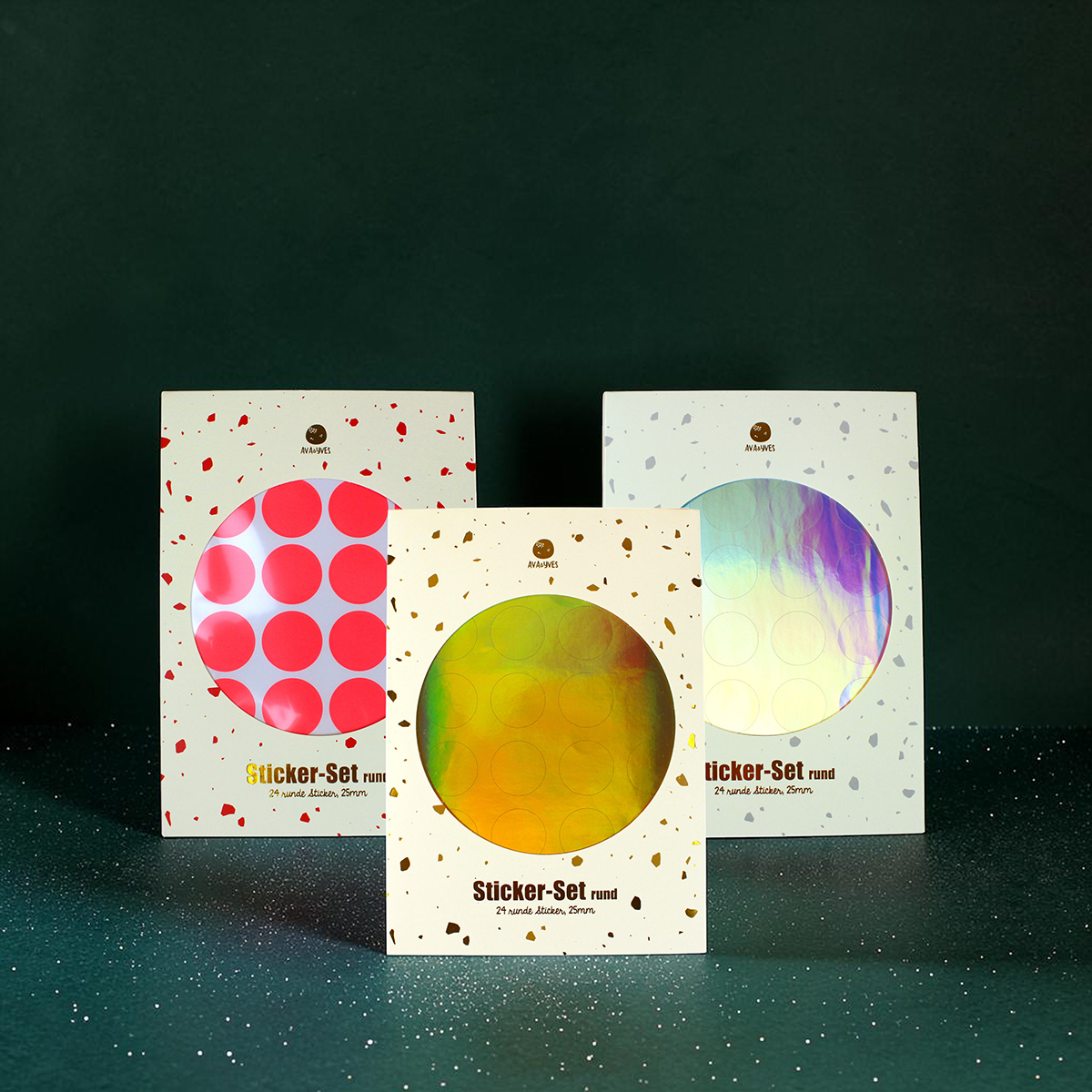Three decorative sticker-pack boxes arranged on a dark green surface, each with a circular holo sticker window and terrazzo pattern; front box shows yellow-green circle, others show pink and iridescent discs.
