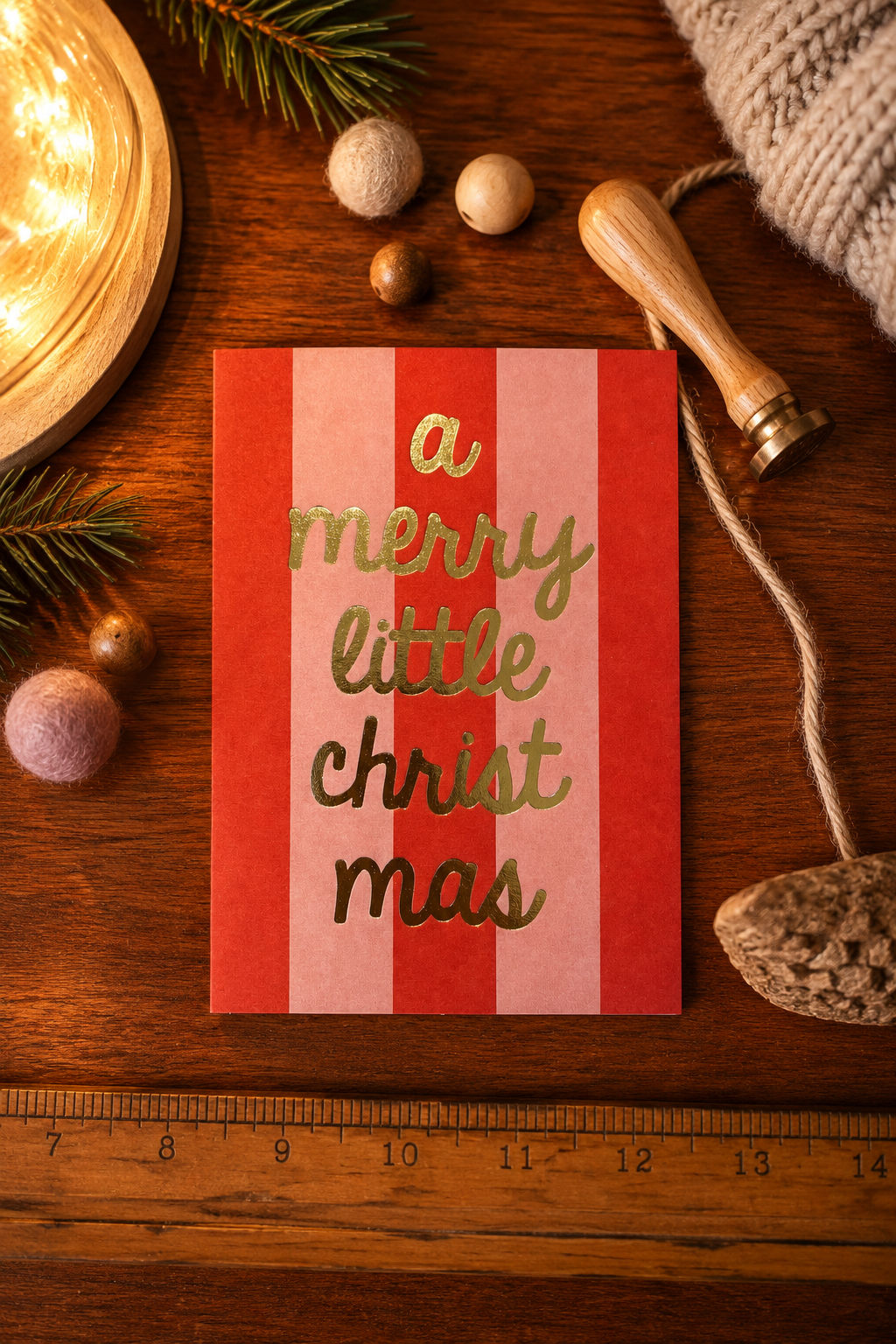 Card with vertical red and pink stripes and gold script that reads 'a merry little christmas' on a wooden surface surrounded by festive props (pine, wool balls, string, ruler).